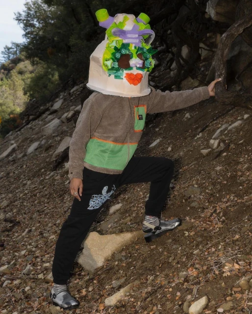 A person wearing a homemade cardboard mask climbing on a hillside with rocks and sparse vegetation, wearing a hooded jacket and pants.