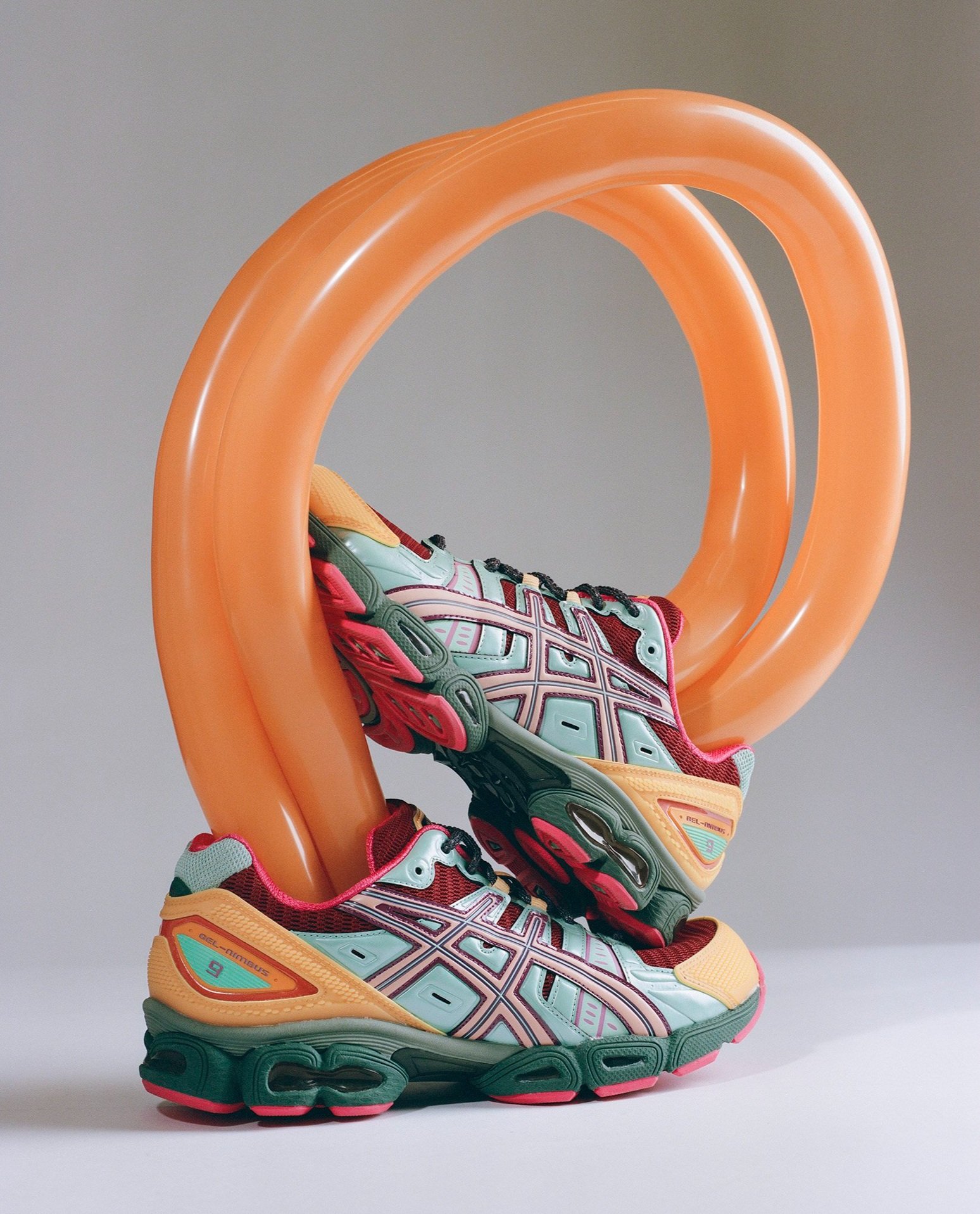 Two athletic shoes are stacked, intertwined with an orange balloon, showcasing a blend of sport and playful design on a plain background.