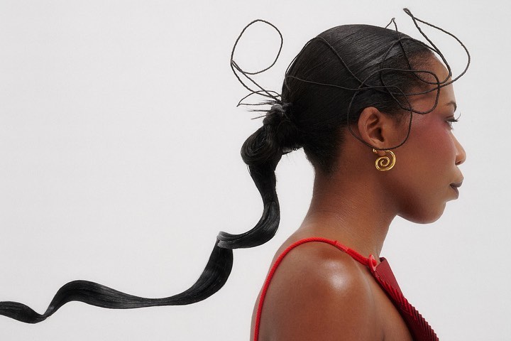 Side profile portrait of Black woman with sleek hair styled with wrapped black string, wearing red top and gold spiral earrings on white.