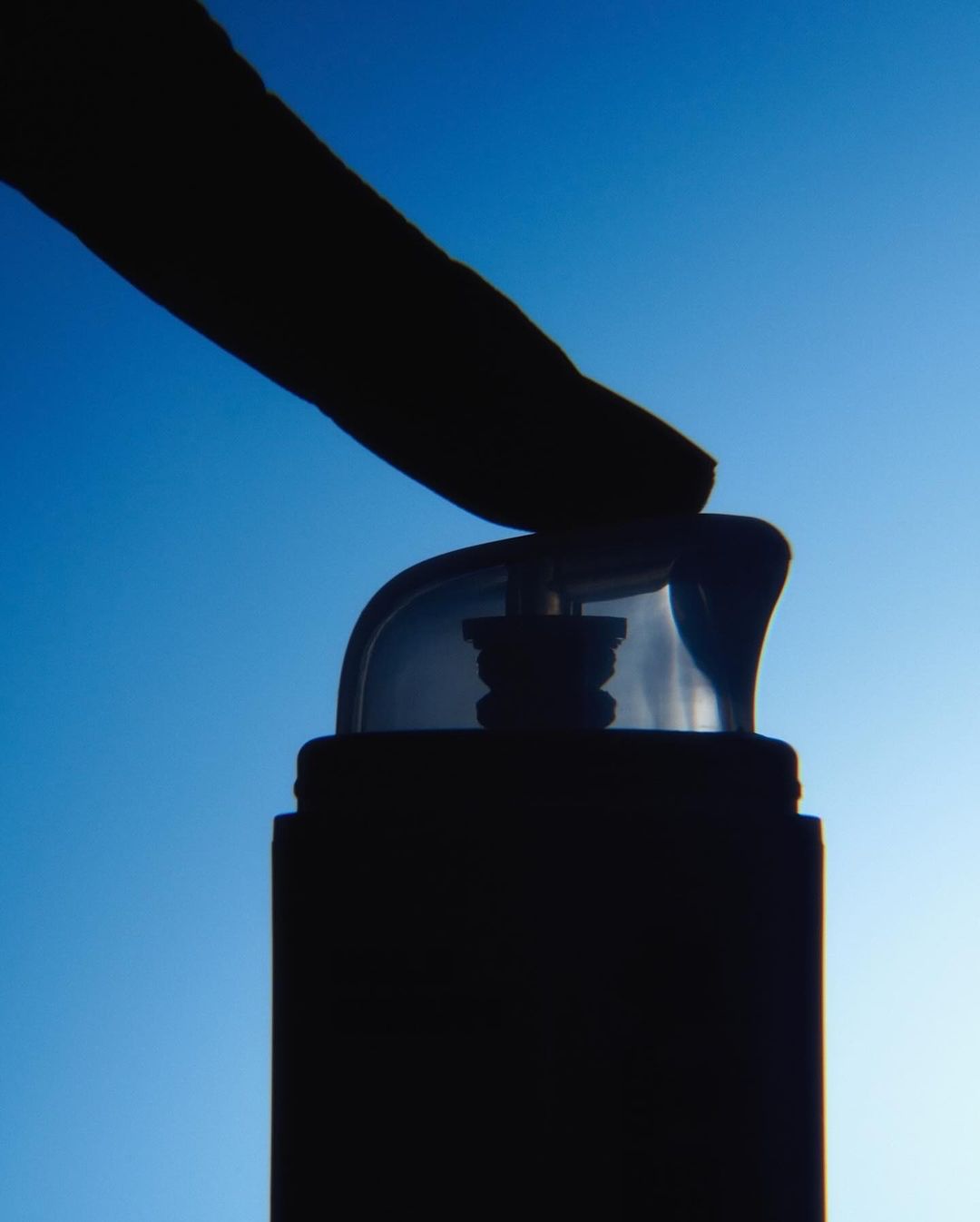 A black silhouette of a finger pressing down on a transparent pump dispenser against a blue sky background, close-up.