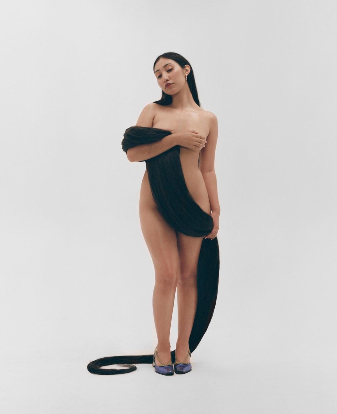 Striking image of an adult female artfully draped with long, dark hair, posing against a plain white backdrop, wearing elegant purple heels.