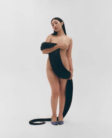 Striking image of an adult female artfully draped with long, dark hair, posing against a plain white backdrop, wearing elegant purple heels.