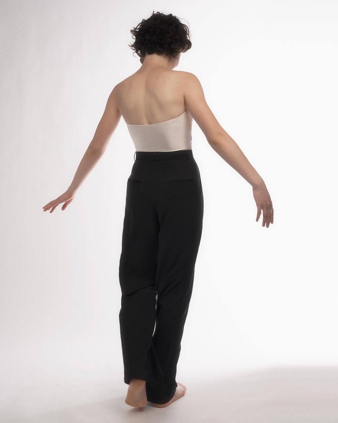 Back view of a slender young adult with short dark hair wearing a beige strapless top and black pants in a white photo studio.