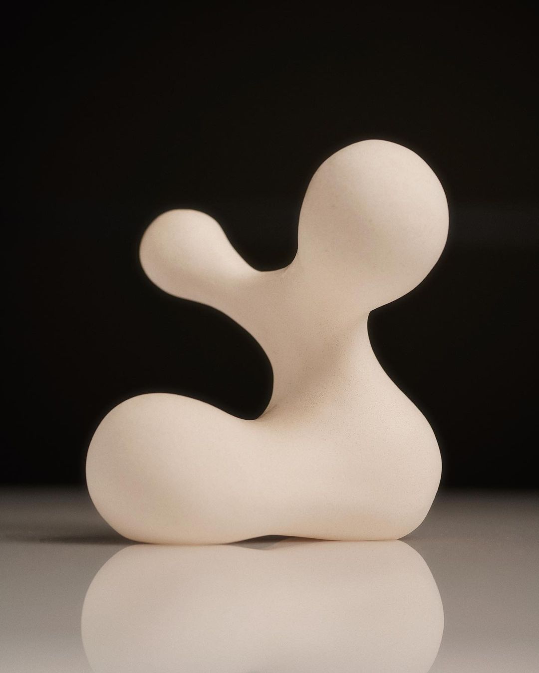 Abstract white sculpture with smooth, rounded forms against a dark background, reflecting on a glossy surface.
