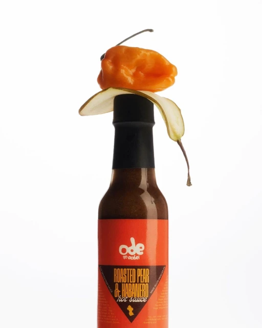 Orange habanero pepper rests atop a bottle of roasted pear and habanero hot sauce, against a plain white background.