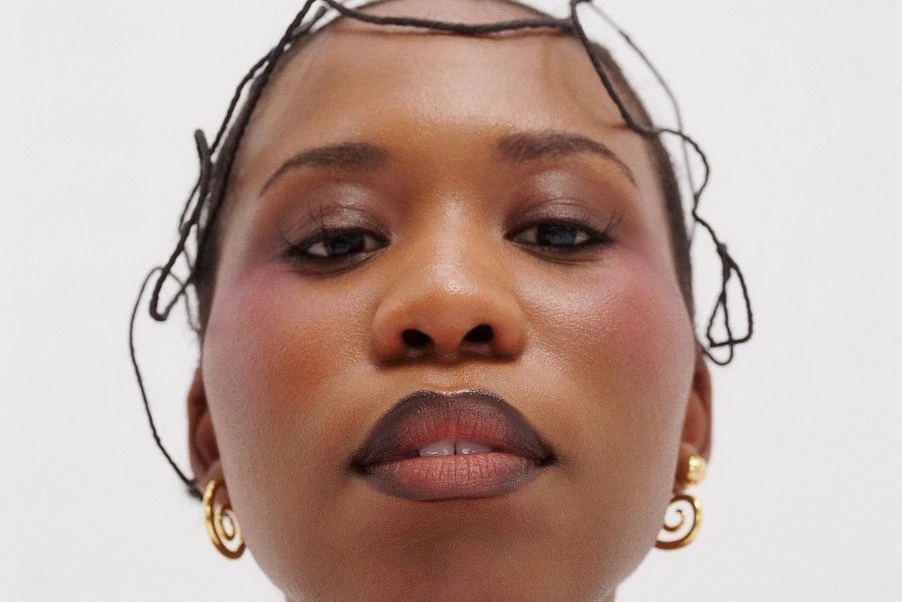 Close-up portrait of a woman with dark hair accessories, wearing gold spiral earrings, and makeup with pink blush on a white background.
