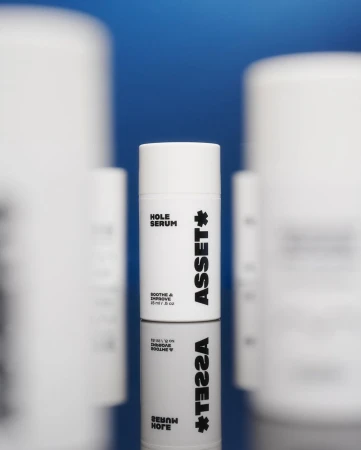 White cylindrical skincare bottle with black text standing upright on a reflective surface against a blue and white backdrop