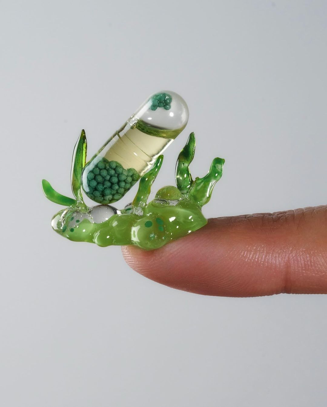 Close-up of a large, clear capsule containing liquid and green beads, being held on a fingertip, set against a plain white background.