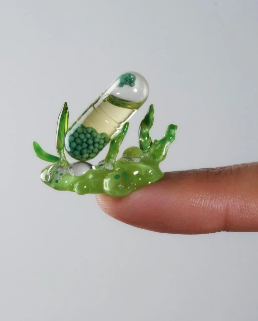 Close-up of a large, clear capsule containing liquid and green beads, being held on a fingertip, set against a plain white background.
