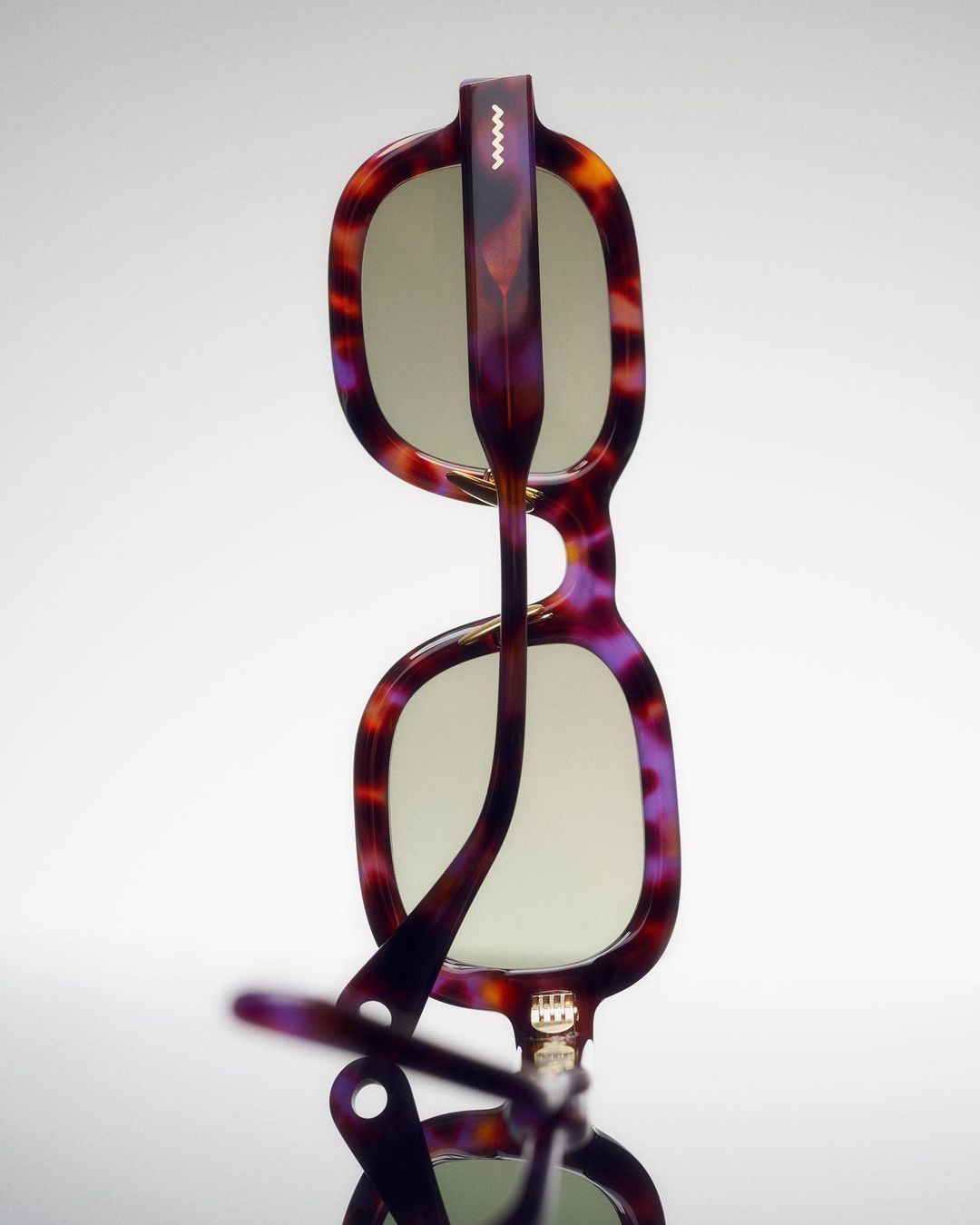 Upside-down view of brown tortoise-shell sunglasses with a purple hue, angled on a reflective surface against a neutral background.