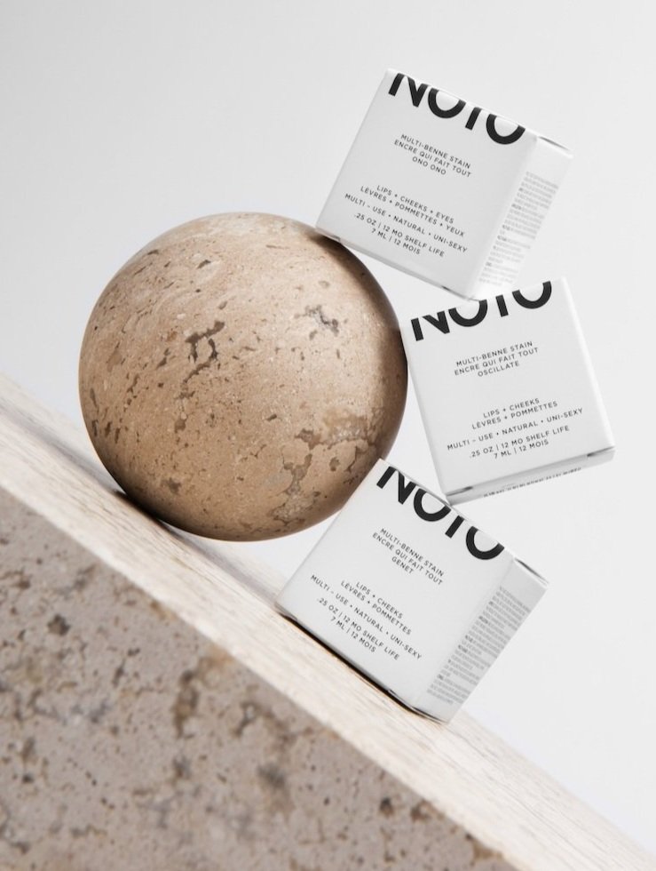 Three NOTO multi-benne stain boxes arranged near a stone ball on a marble surface in a beauty and cosmetic product shot.