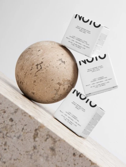 Three NOTO multi-benne stain boxes arranged near a stone ball on a marble surface in a beauty and cosmetic product shot.
