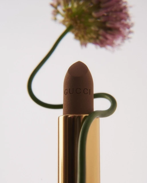 Close-up of brown Gucci lipstick with golden casing, styled with green stems and purple allium flower on a white background.