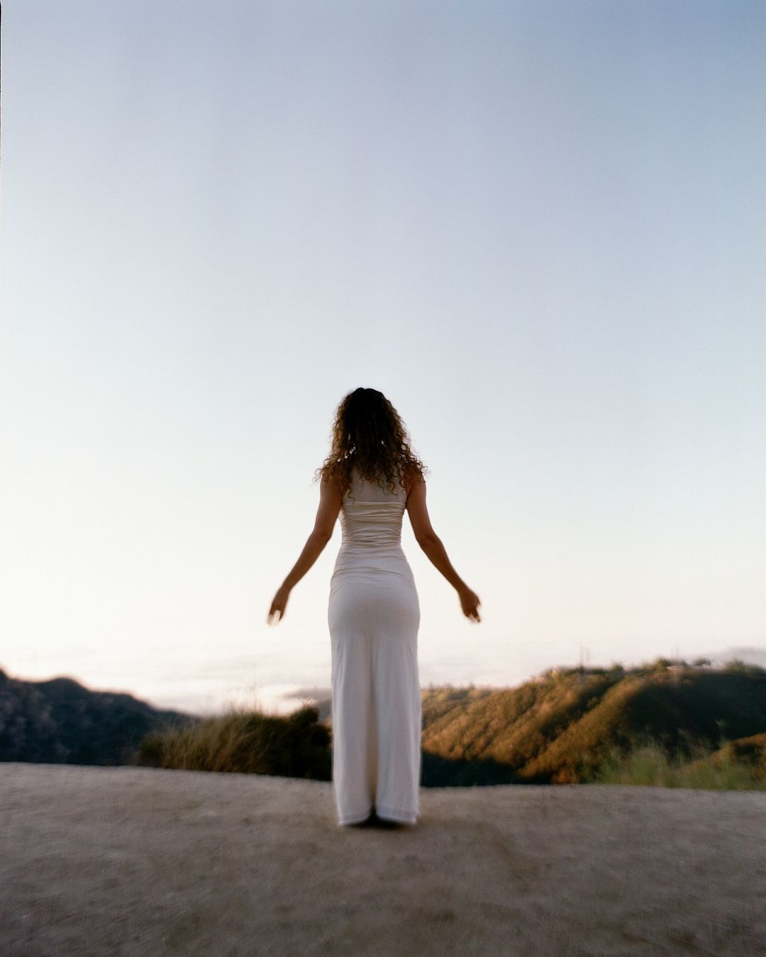 Back view of a curly-haired woman standing with arms outstretched in white dress on a hilltop overlooking a distant valley.