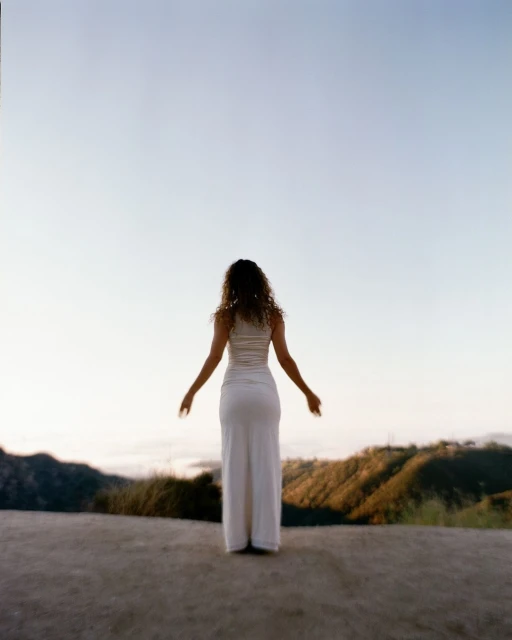 Back view of a curly-haired woman standing with arms outstretched in white dress on a hilltop overlooking a distant valley.