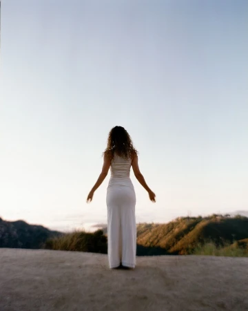Back view of a curly-haired woman standing with arms outstretched in white dress on a hilltop overlooking a distant valley.