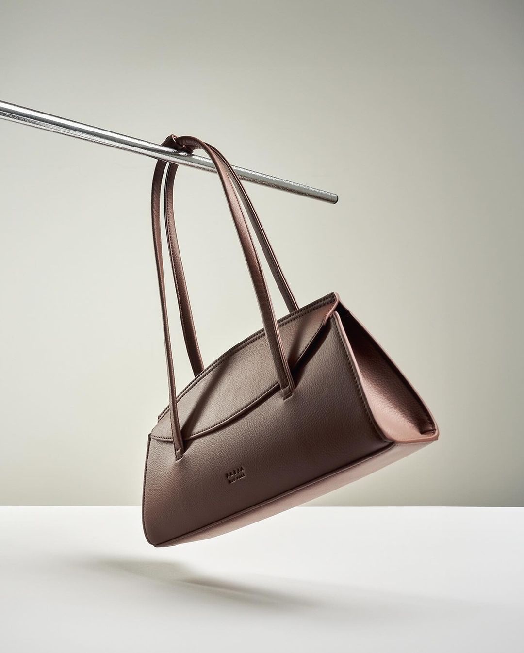 Minimalist studio shot of a brown leather handbag hanging from a silver metal bar against a solid light gray background.
