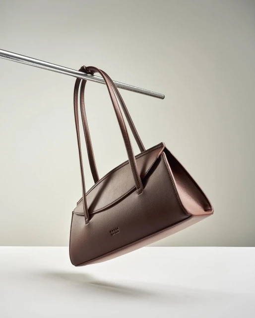 Minimalist studio shot of a brown leather handbag hanging from a silver metal bar against a solid light gray background.