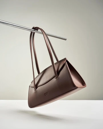 Minimalist studio shot of a brown leather handbag hanging from a silver metal bar against a solid light gray background.