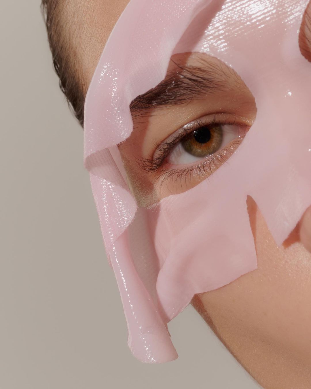 Closeup of person's face covered with translucent pink sheet mask, highlighting eye and skin care treatment on neutral background.