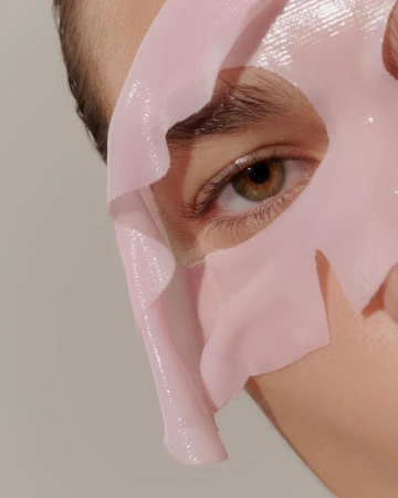 Closeup of person's face covered with translucent pink sheet mask, highlighting eye and skin care treatment on neutral background.