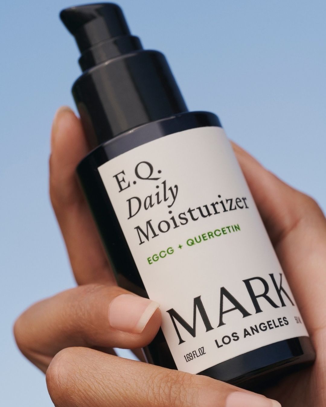 Close-up of a hand holding a black and white bottle of E.Q. Daily Moisturizer by MARK Los Angeles against a sky background.