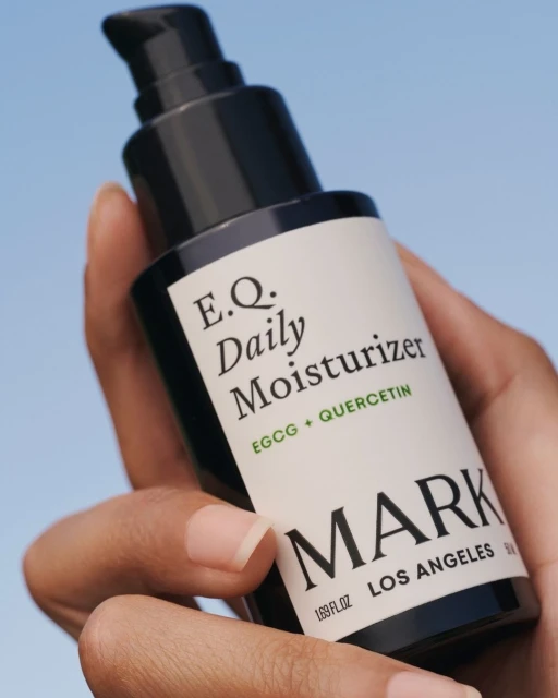 Close-up of a hand holding a black and white bottle of E.Q. Daily Moisturizer by MARK Los Angeles against a sky background.