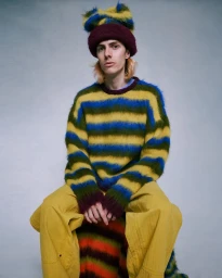 Model wearing a fuzzy striped sweater, layered beanies, and yellow trousers seated against a textured grey background.
