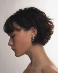 Profile portrait of a woman with dark short wavy hair and multiple silver earrings against a plain white background
