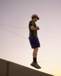 A man wearing a green t-shirt and purple shorts jumping rope outdoors at sunset against a clear sky.