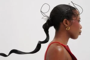 Profile portrait of a black woman wearing avant-garde sculptural hair styling and gold swirl earrings against a white background.
