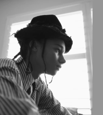 Monochrome portrait of a young person in profile wearing a dark fedora and striped button-down shirt by a window.