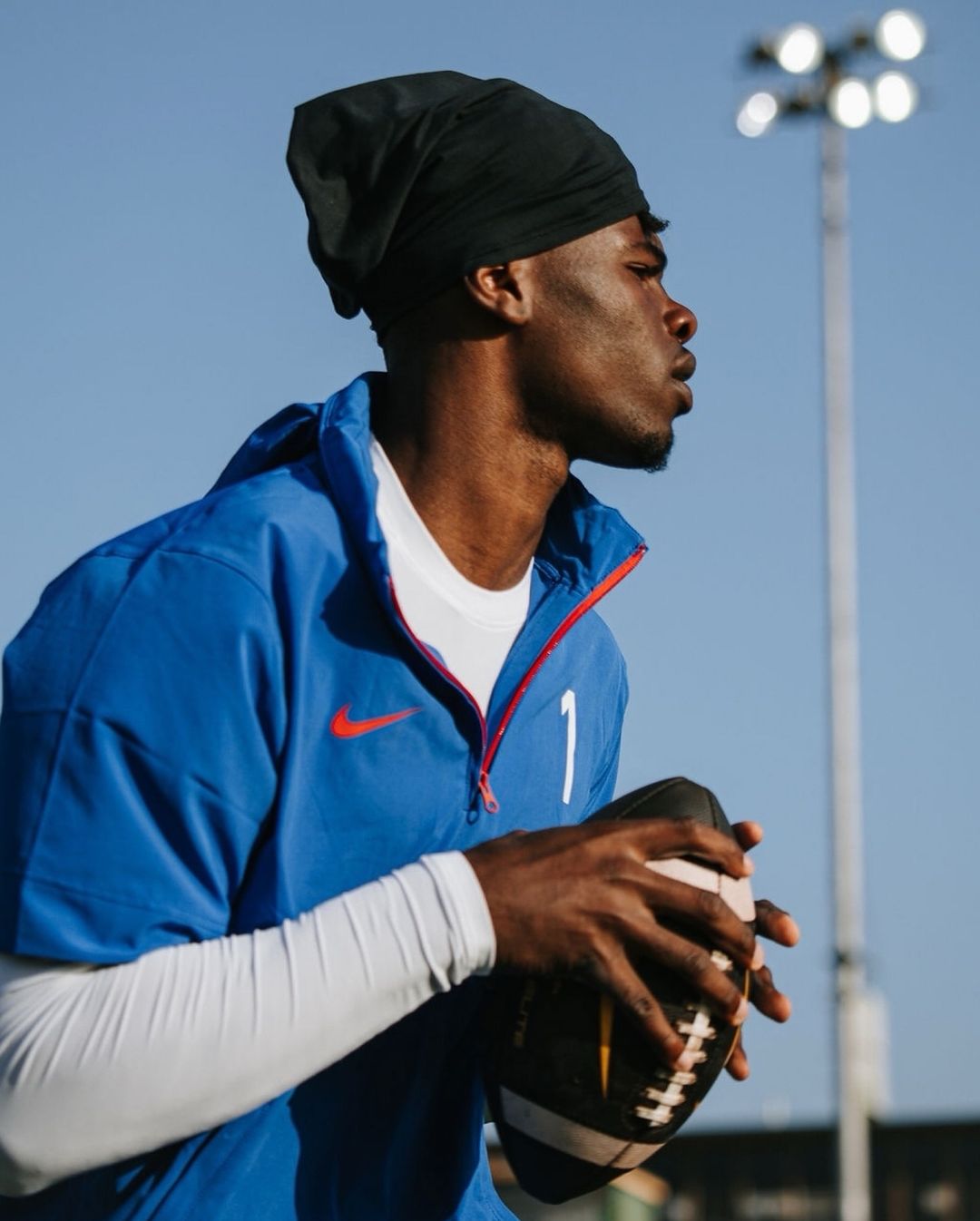 Athlete wearing blue sports jersey and black head covering holding a football outdoors under clear blue sky.