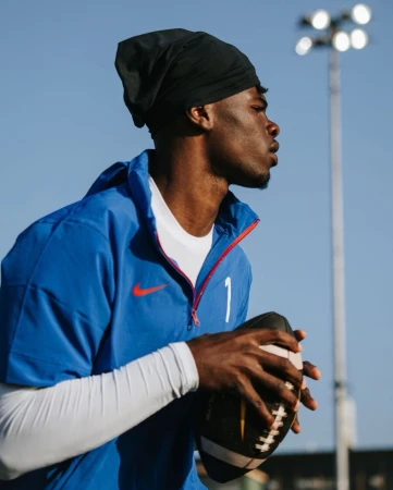 Athlete wearing blue sports jersey and black head covering holding a football outdoors under clear blue sky.