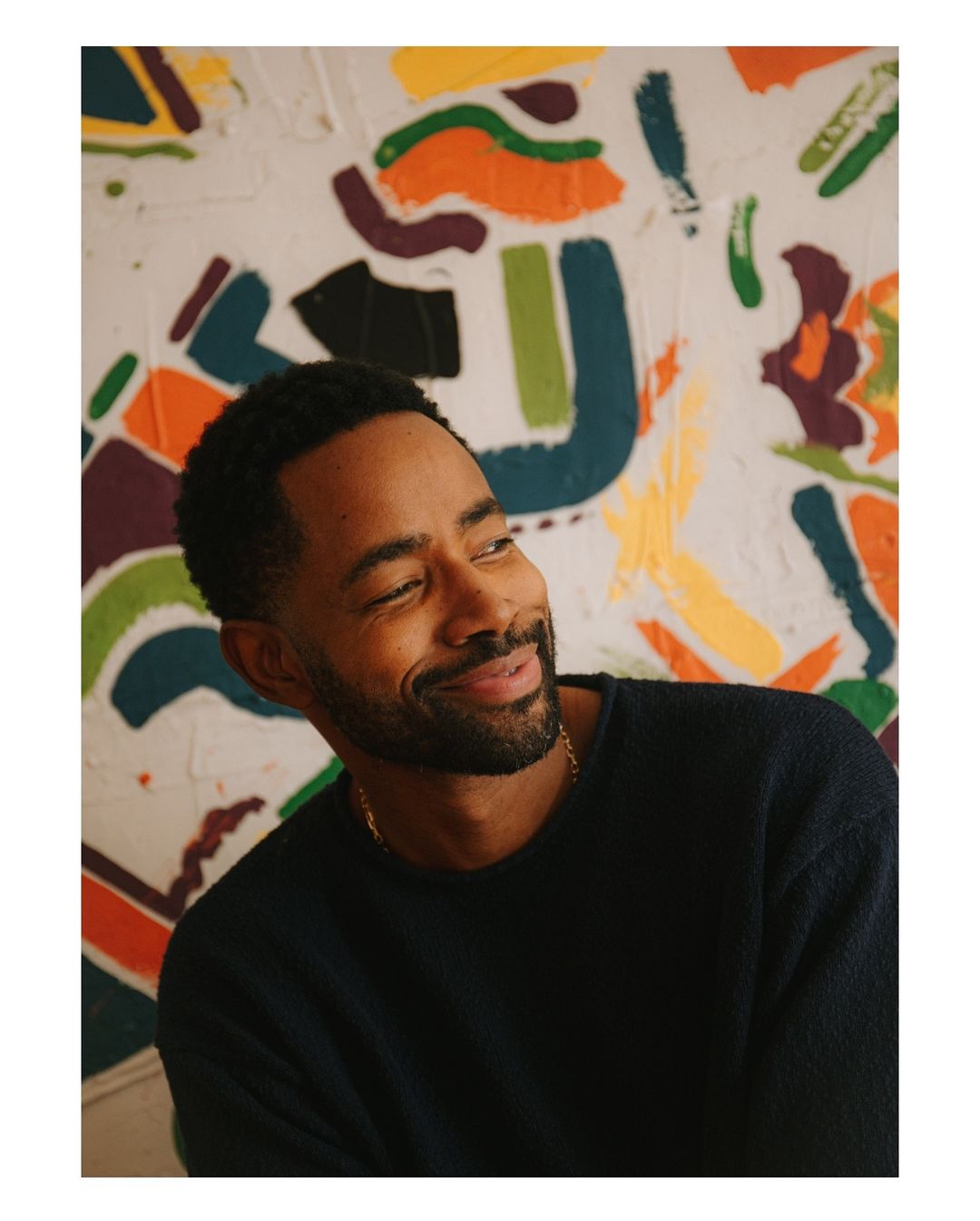Smiling Black man with a beard in a navy sweater, posing indoors in front of a white wall with abstract multicolored art.