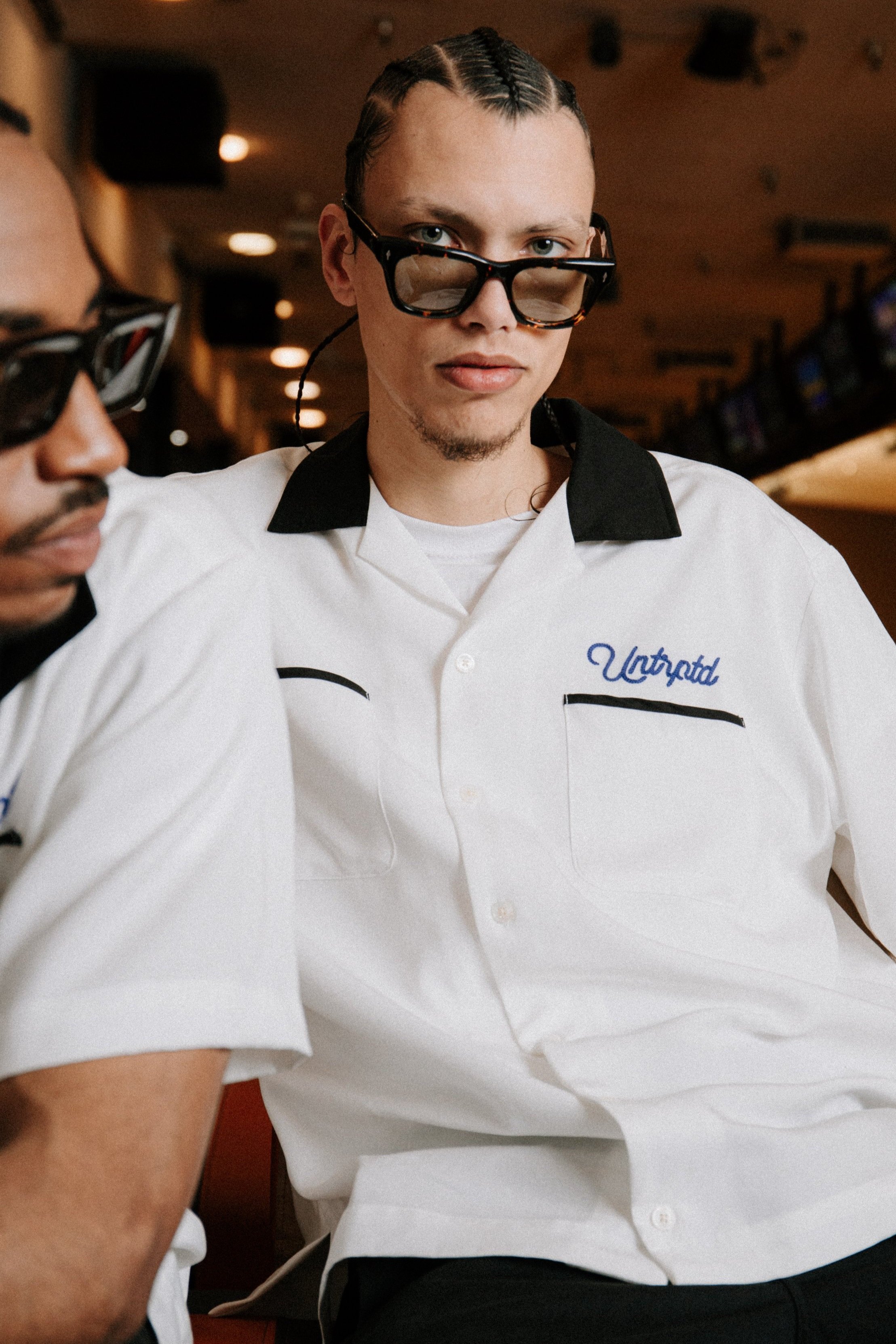 Two men wear retro bowling shirts and sunglasses indoors, one facing the camera with braided hair, stylish and fashionable.
