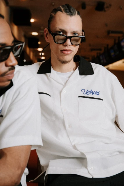 Two men wear retro bowling shirts and sunglasses indoors, one facing the camera with braided hair, stylish and fashionable.