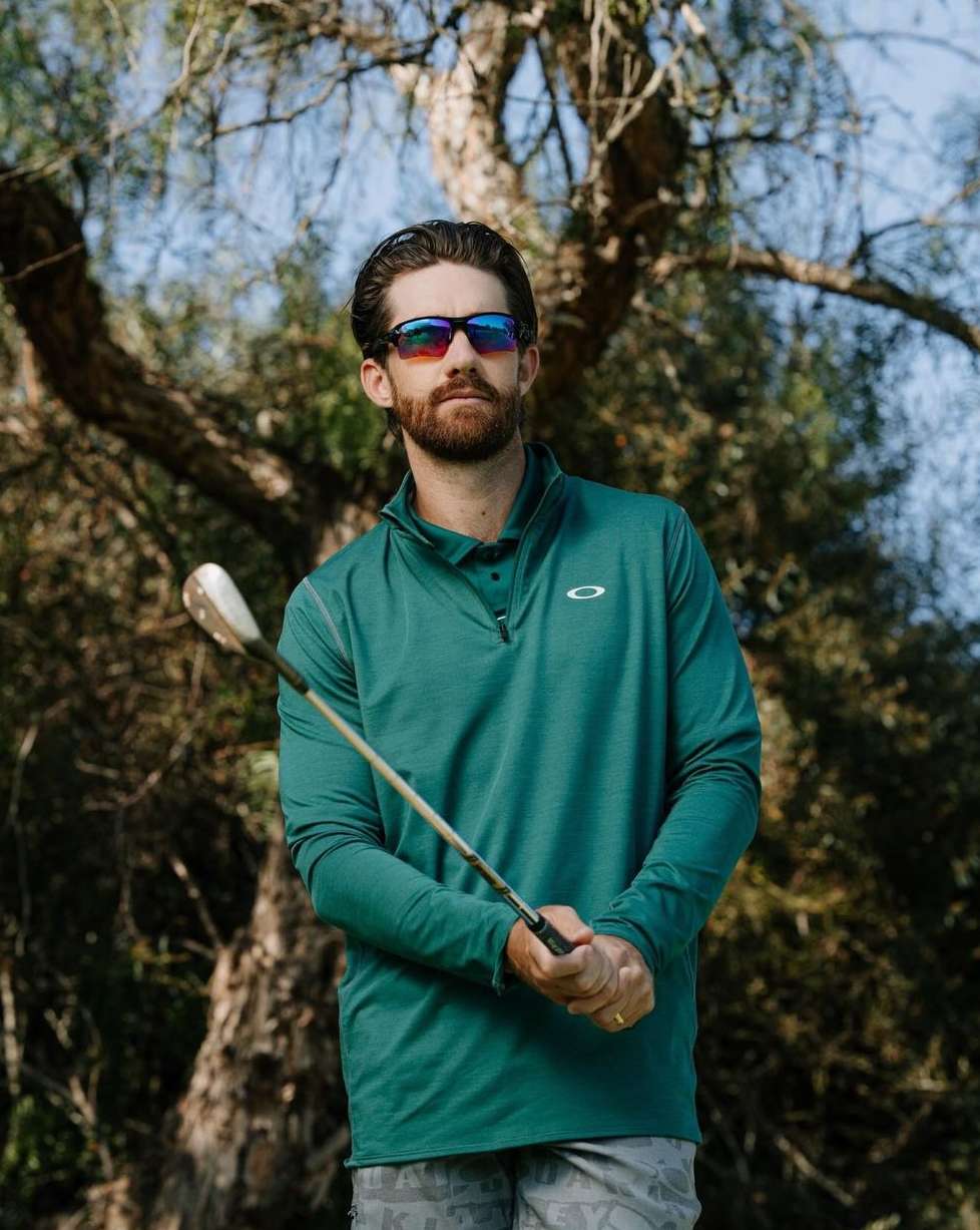 Portrait of a man in athletic wear outdoors holding a golf club, with trees and foliage in the background under a blue sky.