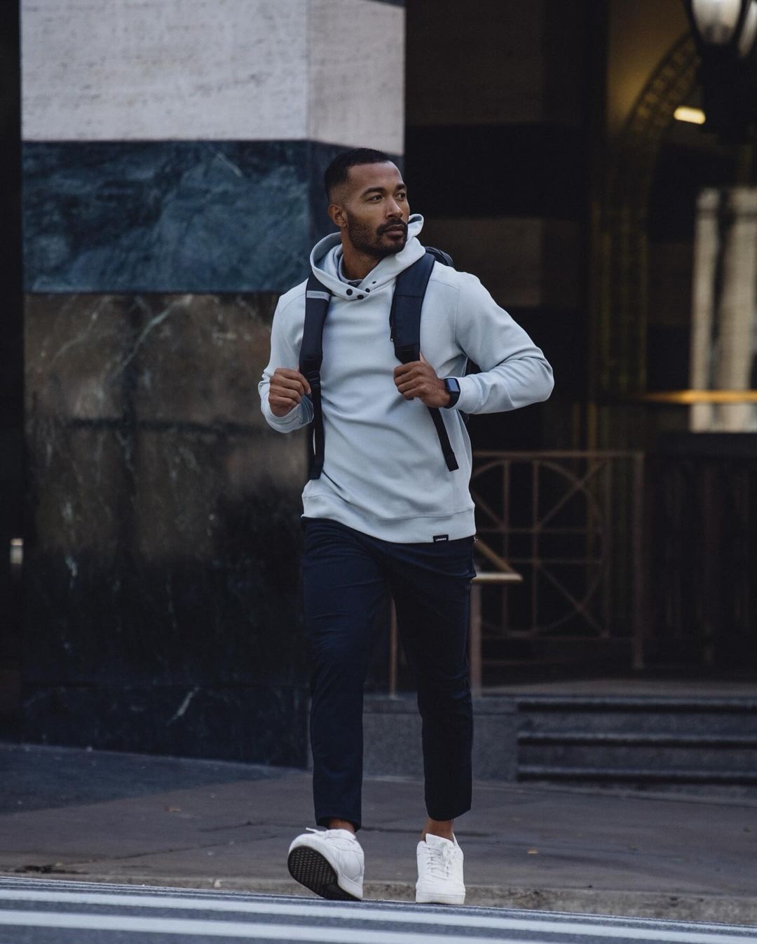 Full-body shot of an adult Black male in a hoodie and backpack crossing a city street on a crosswalk with buildings in the background.