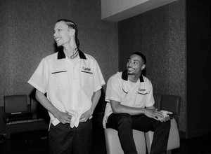 Two men in bowling shirts in black and white. One man is smiling and looking off to the side; the other is seated and also smiling.