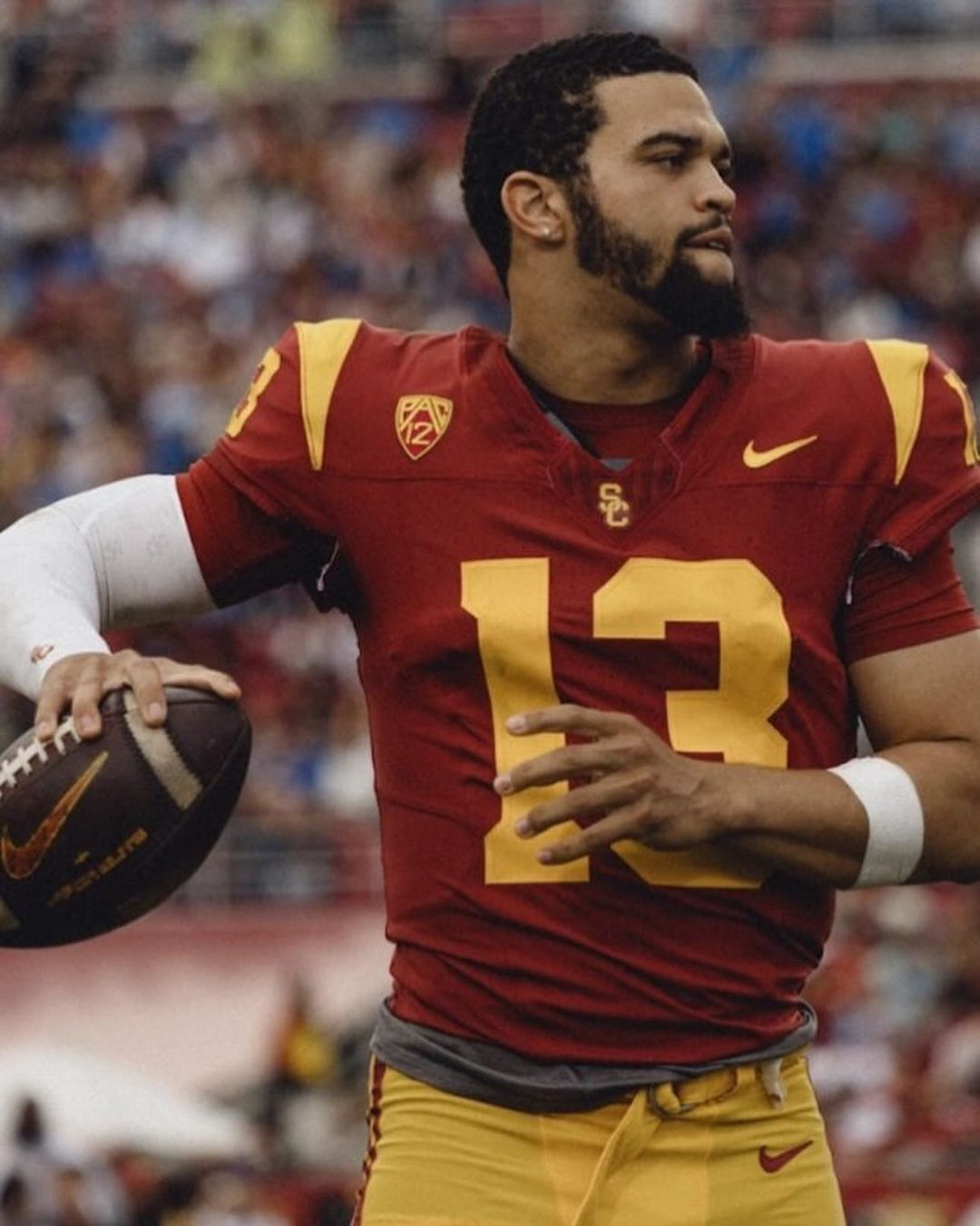 Football player in maroon and gold uniform holding a football, looking left, wearing gold pants with stadium background.