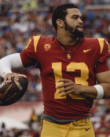Football player in maroon and gold uniform holding a football, looking left, wearing gold pants with stadium background.