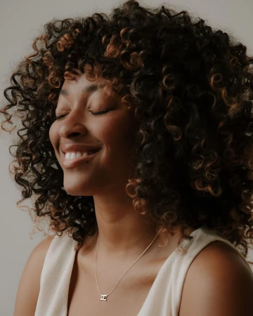 Portrait of a Black woman with voluminous curly hair smiling with eyes closed, wearing a cream top and delicate necklace.