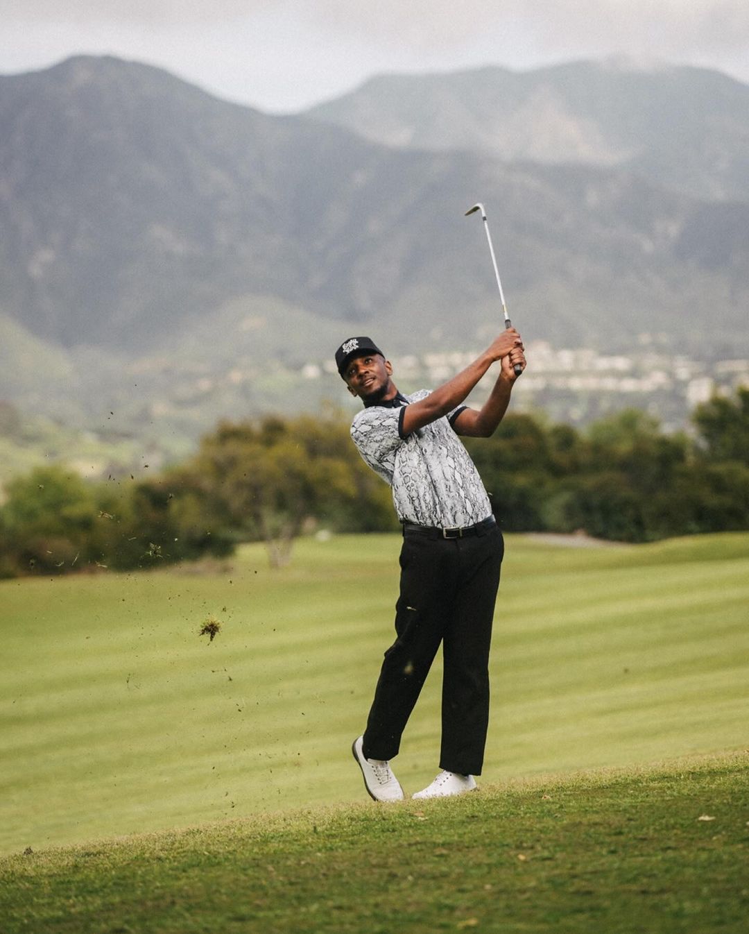 A golfer in mid-swing on a lush green golf course with mountains in the background, wearing black pants and a patterned shirt.