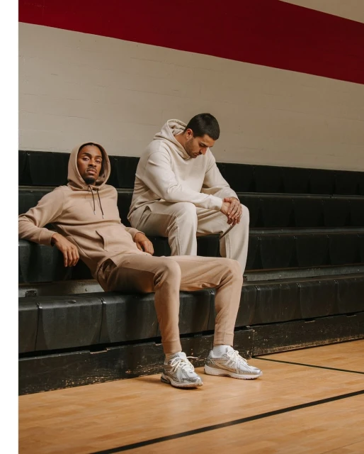 Two men in beige hoodies and sweatpants sitting on black bleachers. One is looking down, the other is looking at the camera.