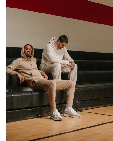 Two men in beige hoodies and sweatpants sitting on black bleachers. One is looking down, the other is looking at the camera.