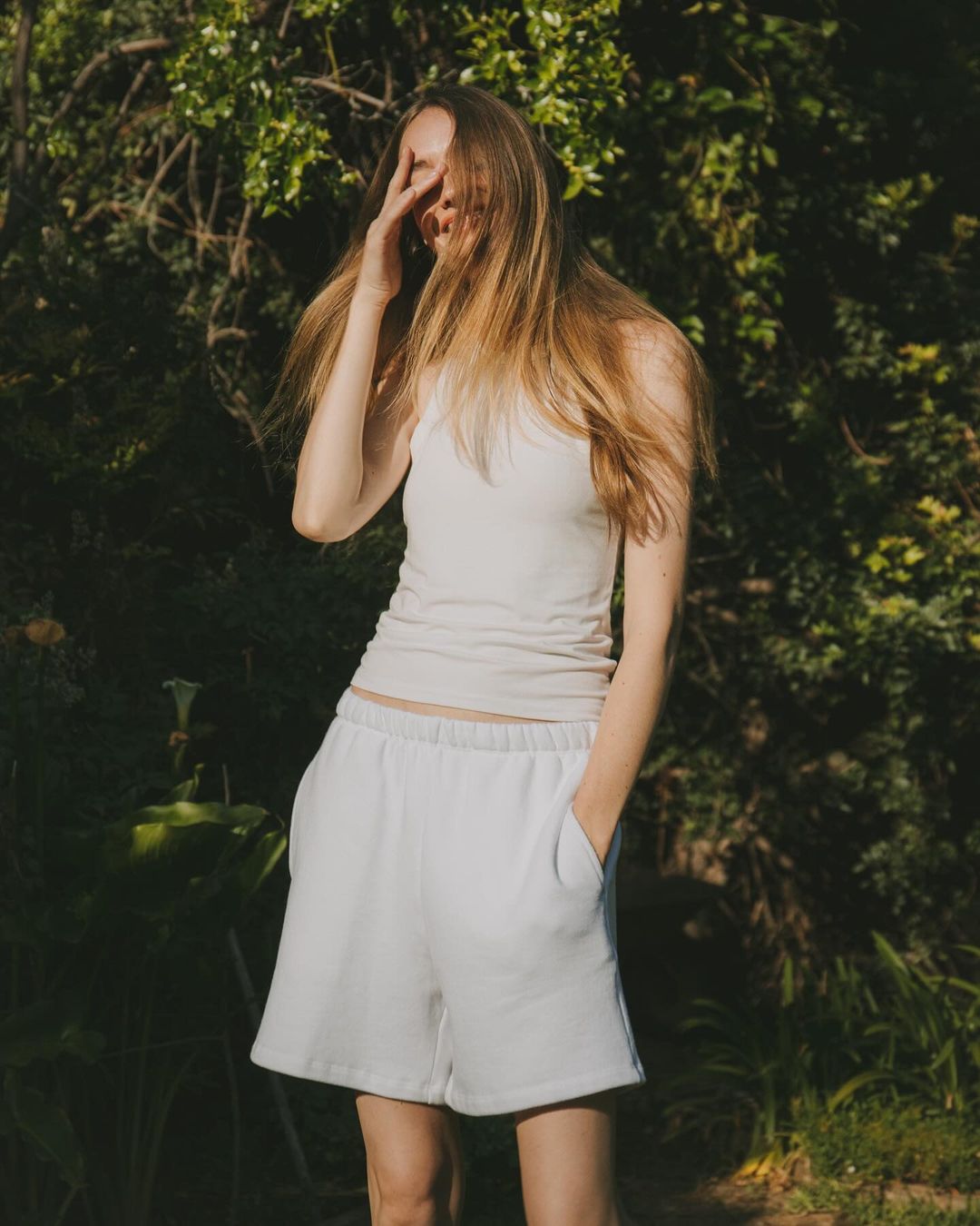 Young woman in casual summer clothing standing outdoors in a natural setting, shielding her eyes from the sunlight on a bright day.