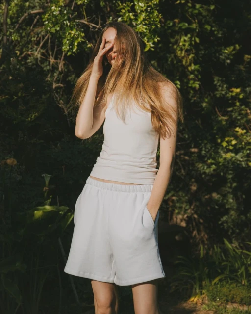 Young woman in casual summer clothing standing outdoors in a natural setting, shielding her eyes from the sunlight on a bright day.