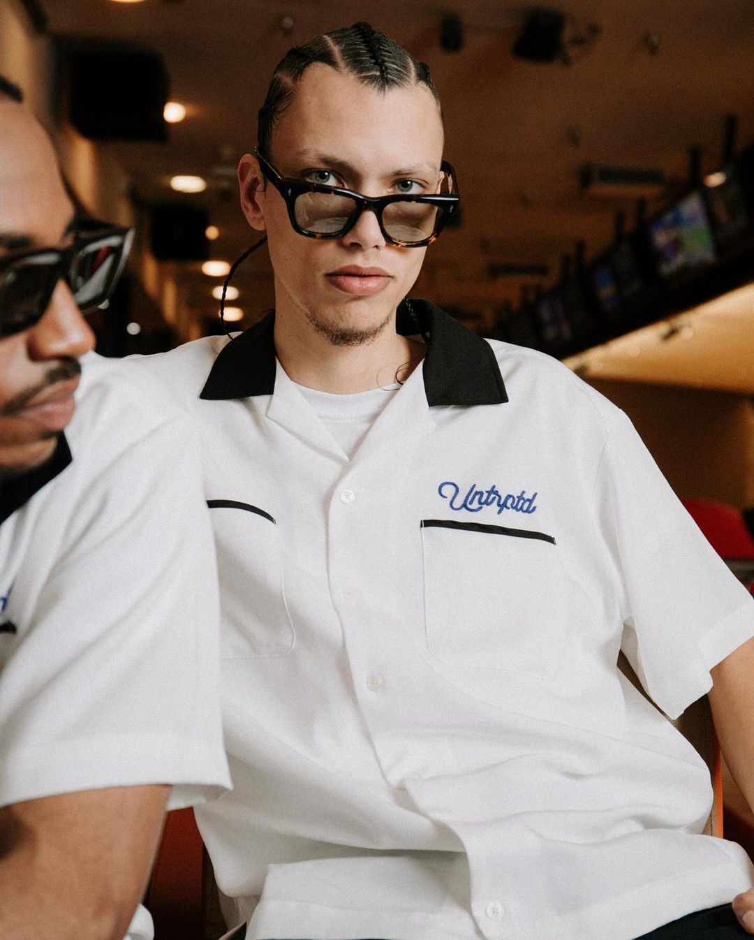 Two men pose indoors wearing white shirts with black collars and retro sunglasses. Bowling alley in background, medium shot.
