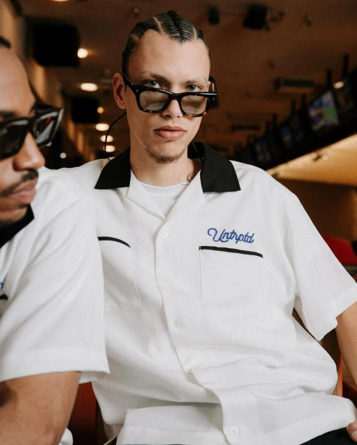 Two men pose indoors wearing white shirts with black collars and retro sunglasses. Bowling alley in background, medium shot.